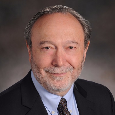 A professional headshot of a middle-aged man with a slight smile, wearing a dark suit and tie, against a neutral gray background.