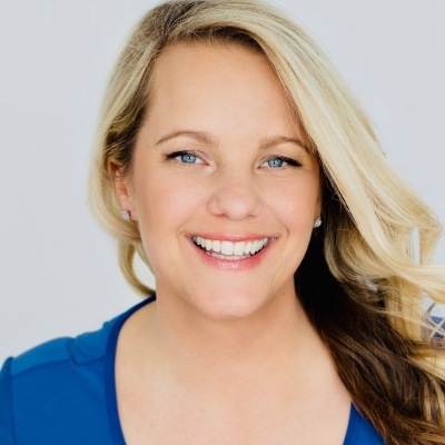 A smiling woman with bright blue eyes and blond hair, dressed in a blue top and showing a confident, friendly demeanor.
