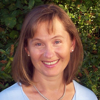 A smiling woman with short brown hair wearing a blue top and a silver necklace, with a background of green foliage.