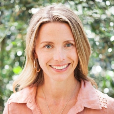 Portrait of a smiling woman with blond hair, wearing a light pink blouse against a natural, leafy background.