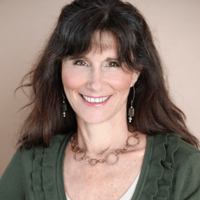 A smiling woman with long, dark hair and bangs, wearing earrings, a green top, and a brown necklace, posing for a portrait.