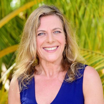 A smiling woman with long wavy hair wearing a blue sleeveless top, posing in front of a lush green background with palm fronds.