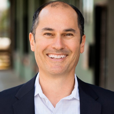 A confident professional man smiling at the camera, dressed in business attire with a blue shirt and dark jacket.