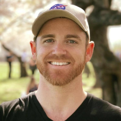 A smiling man with a beard wearing a baseball cap outdoors on a sunny day.