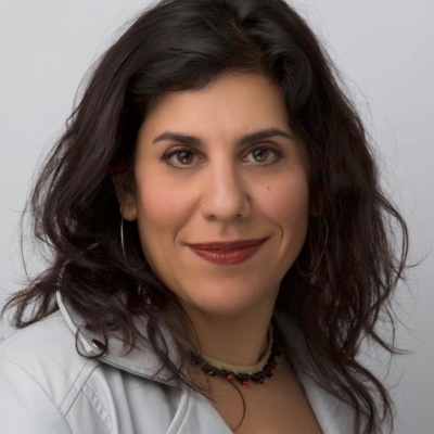 Professional headshot of a woman with a friendly smile, featuring her dark wavy hair and a simple necklace, against a neutral background.