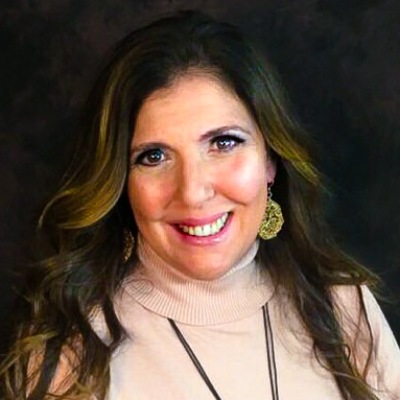 A smiling woman with long wavy hair wearing a turtleneck sweater and statement earrings against a dark backdrop.
