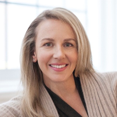 A professional headshot of a smiling woman with blonde hair, dressed in a beige cardigan and a black top, conveying confidence and approachability.