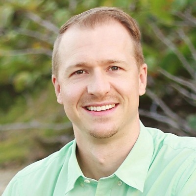 A smiling man in a light green polo shirt posing for a friendly, approachable portrait with a natural backdrop.