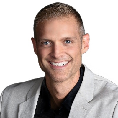 A smiling man with short hair, wearing a gray blazer over a black shirt, set against a plain background.
