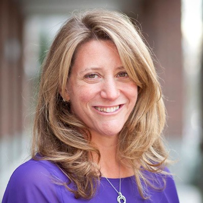 A smiling woman with shoulder-length blonde hair wearing a purple blouse and a necklace, posing for a friendly portrait outdoors.