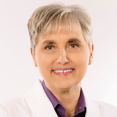 A professional portrait of a smiling woman with short gray hair, wearing a white lab coat over a purple blouse, exuding confidence and experience.