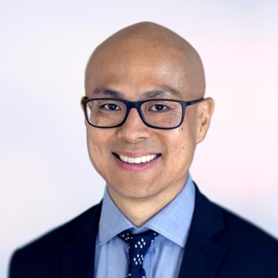 A professional portrait of a man with glasses, smiling and wearing a blue shirt with a tie.