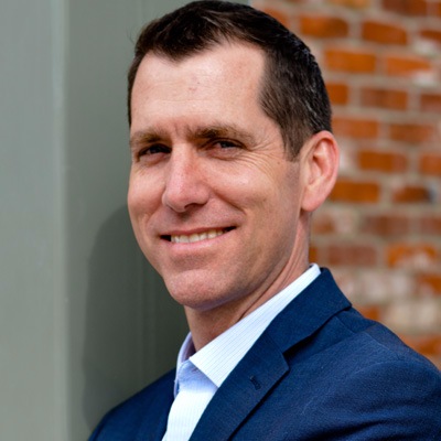 A professional man in a suit smiling with confidence against a brick wall background.
