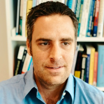 A professional man with dark hair and a blue shirt posing for a portrait in an office setting, featuring bookshelves in the background.