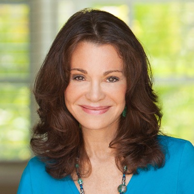 A professional woman with brown hair smiling confidently at the camera, wearing a blue top and a necklace with a blurred natural backdrop.
