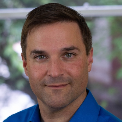 A confident professional in a blue shirt poses for a headshot with a slight smile and a backdrop of greenery.