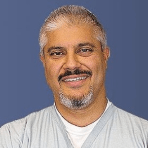 A man with salt-and-pepper hair and a goatee, wearing a light grey t-shirt, is smiling against a blue background.