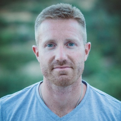A portrait of a man with short blonde hair and a beard, wearing a gray t-shirt, set against a blurred natural backdrop. he has a serious expression on his face as he looks directly at the camera.