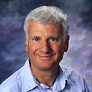 Smiling man with gray hair posing for a professional portrait against a mottled blue background.