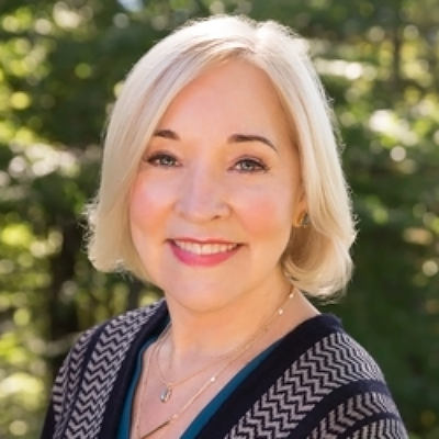 A smiling woman with short blonde hair, wearing a black and white patterned top, posing for a portrait with a backdrop of lush green foliage.