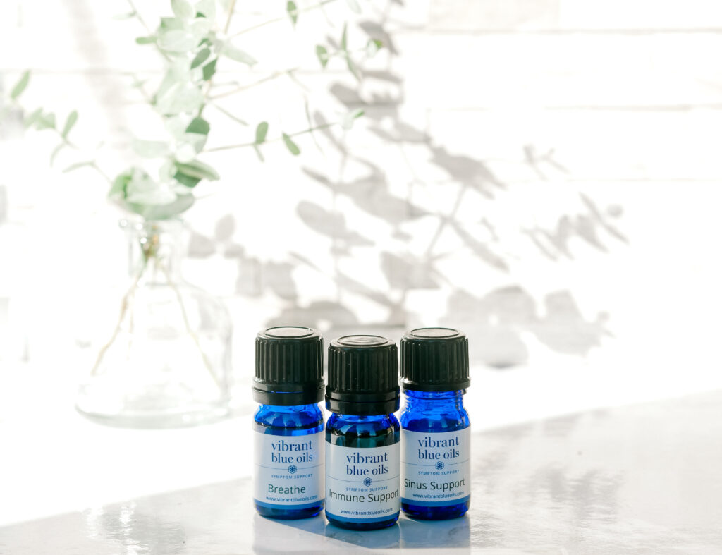 A trio of blue bottles of vibrant blue oils on a reflective surface, each labeled for a different purpose—breathe, immune support, sinus support—against a backdrop of soft natural light and delicate foliage.
