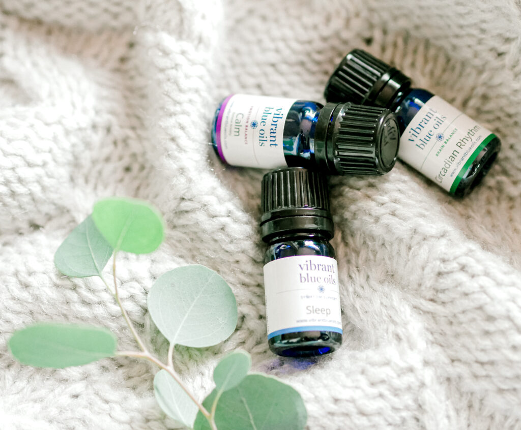 Three small bottles of essential oils with labels indicating different types, placed on a soft, textured white blanket, accompanied by a few delicate green leaves for a touch of natural elegance.