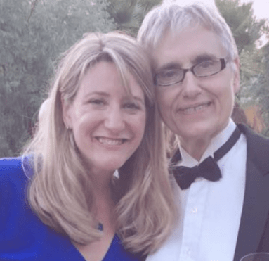 A smiling duo posing for a photo, the woman with long blonde hair and the man sporting glasses and a bow tie. they're dressed for a formal occasion amidst a backdrop of greenery.